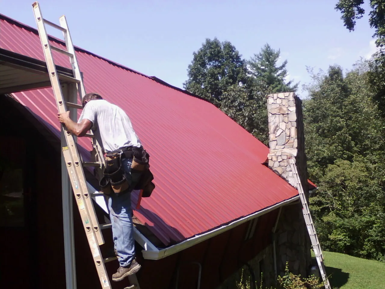 Roofer on a ladder beside a red metal roof and stone chimney — Soffit & Fascia Repair by Roofing Brothers in Van Hornesville